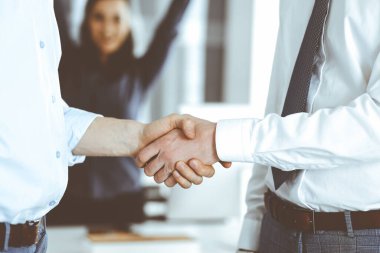 Ofiste iki işadamı el sıkışıyor, yakın plan. Mutlu ve heyecanlı iş kadını arka planda elini kaldırıyor. İş adamları konsepti