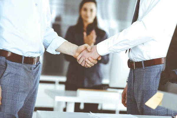 Ofiste iki işadamı el sıkışıyor, yakın plan. Mutlu ve heyecanlı iş kadını arka planda elini kaldırıyor. İş adamları konsepti