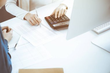 Muhasebeci mali durumu kontrol ediyor ya da vergi formları için hesap makinesi geliri ile sayıyor, eller yakın çekim. İş kadını ofisteki masada iş arkadaşıyla oturuyor ve çalışıyor. Denetim kavramı