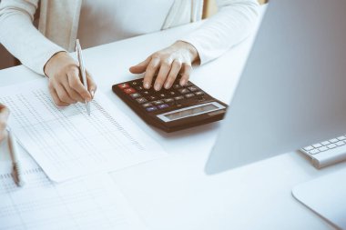 Muhasebeci mali durumu kontrol ediyor ya da vergi formları için hesap makinesi geliri ile sayıyor, eller yakın çekim. İş kadını ofisteki masada iş arkadaşıyla oturuyor ve çalışıyor. Denetim kavramı