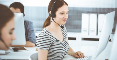 İnternette müşterilerle konuşurken kulaklık ve bilgisayar kullanan sıradan giyinmiş genç bir kadın. Bir grup operatör iş başında. Çağrı merkezi, iş konsepti