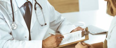 Doktor ve hasta klinikteki masada otururken tıbbi muayene sonuçlarını tartışıyorlar, yakın plan. Genç bir kadının ilaç geçmişini doldurmak için pano kullanan bir erkek doktor.