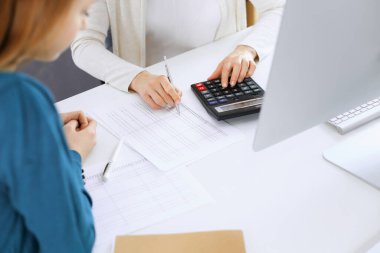 Muhasebeci mali durumu kontrol ediyor ya da vergi formları için hesap makinesi geliri ile sayıyor, eller yakın çekim. İş kadını ofisteki masada iş arkadaşıyla oturuyor ve çalışıyor. Denetim kavramı