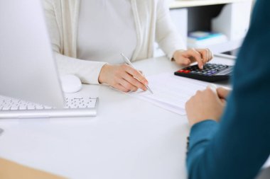 Muhasebeci mali durumu kontrol ediyor ya da vergi formları için hesap makinesi geliri ile sayıyor, eller yakın çekim. İş kadını ofisteki masada iş arkadaşıyla oturuyor ve çalışıyor. Denetim kavramı