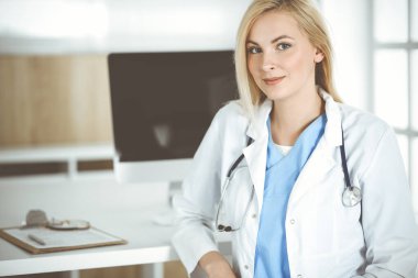 Hastanedeki ya da klinikteki masa başında oturan kadın doktor. Sarışın, neşeli doktor hastalara yardım etmeye hazır.