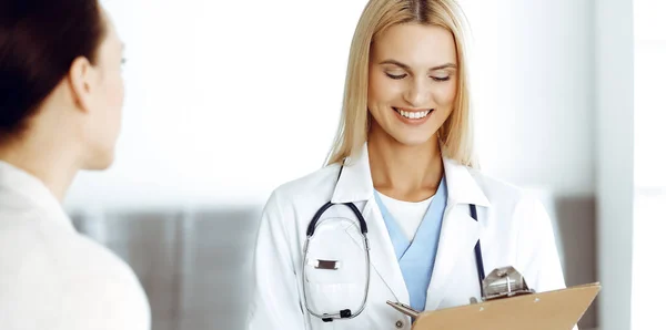 Woman-doctor at work in hospital is happy to consult female patient. Blonde physician checks medical history record and exam results while using clipboard. Medicine concept