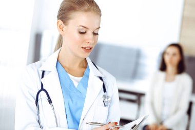 Kadın - doktor hastanın arka planında klinikte tıbbi geçmiş kayıtlarını doldurmak için pano kullanıyor. Tıp konsepti