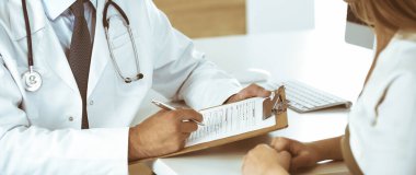 Doktor ve hasta klinikteki masada otururken tıbbi muayene sonuçlarını tartışıyorlar, yakın plan. Genç bir kadının ilaç geçmişini doldurmak için pano kullanan bir erkek doktor.