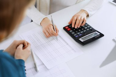 Muhasebeci mali durumu kontrol ediyor ya da vergi formları için hesap makinesi geliri ile sayıyor, eller yakın çekim. İş kadını ofisteki masada iş arkadaşıyla oturuyor ve çalışıyor. Denetim kavramı