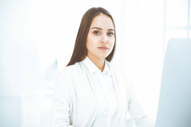 Genç, neşeli bir kadın bilgisayarla masada oturuyor ve beyaz renkli ofisteki kameraya bakıyor. Öğrenci kız ya da iş kadınına benziyor. Vesikalık