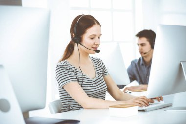 İnternette müşterilerle konuşurken kulaklık ve bilgisayar kullanan sıradan giyinmiş genç bir kadın. Bir grup operatör iş başında. Çağrı merkezi, iş konsepti