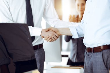 Güneşli ofiste el sıkışan iki işadamı, yakın plan. Mutlu ve heyecanlı iş kadını arka planda elini kaldırıyor. İş adamları konsepti