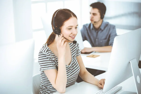 Casual dressed young woman using headset and computer while talking with customers online. Group of operators at work. Call center, business concept