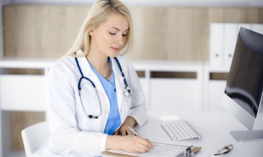 Hastanedeki ya da klinikteki masa başında oturan kadın doktor. Sarışın, neşeli doktor ilaç geçmişi formunu dolduruyor.