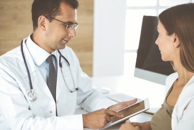 Doktor ve hasta klinikteki masada otururken tıbbi muayene sonuçlarını tartışıyorlar. Genç bir kadının ilaç geçmişini doldurmak için tablet bilgisayar kullanan bir erkek doktor. Tıptaki veriler