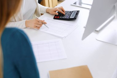 Muhasebeci mali durumu kontrol ediyor ya da vergi formları için hesap makinesi geliri ile sayıyor, eller yakın çekim. İş kadını ofisteki masada iş arkadaşıyla oturuyor ve çalışıyor. Denetim kavramı