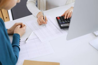 Muhasebeci mali durumu kontrol ediyor ya da vergi formları için hesap makinesi geliri ile sayıyor, eller yakın çekim. İş kadını ofisteki masada iş arkadaşıyla oturuyor ve çalışıyor. Denetim kavramı