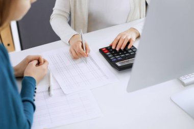 Muhasebeci mali durumu kontrol ediyor ya da vergi formları için hesap makinesi geliri ile sayıyor, eller yakın çekim. İş kadını ofisteki masada iş arkadaşıyla oturuyor ve çalışıyor. Denetim kavramı