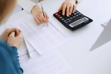 Muhasebeci mali durumu kontrol ediyor ya da vergi formları için hesap makinesi geliri ile sayıyor, eller yakın çekim. İş kadını ofisteki masada iş arkadaşıyla oturuyor ve çalışıyor. Denetim kavramı