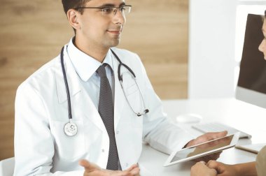 Doktor ve hasta klinikteki masada otururken tıbbi muayene sonuçlarını tartışıyorlar. Genç bir kadının ilaç geçmişini doldurmak için tablet bilgisayar kullanan bir erkek doktor. Tıptaki veriler