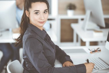 Modern ofisteki işyerinde bilgisayar kullanan bir iş kadını. Esmer sekreter ya da kadın avukat kameraya bakıyor ve mutlu gülümsüyor. Zevk ve başarı için çalışmak