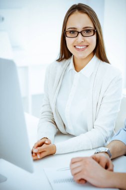 Genç, neşeli bir kadın bilgisayarla masada oturuyor ve beyaz renkli ofisteki kameraya bakıyor. Öğrenci kız ya da iş kadınına benziyor. Vesikalık