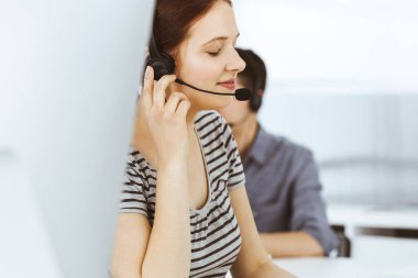 İnternette müşterilerle konuşurken kulaklık ve bilgisayar kullanan sıradan giyinmiş genç bir kadın. Bir grup operatör iş başında. Çağrı merkezi, iş konsepti