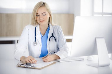 Hastanedeki ya da klinikteki masa başında oturan kadın doktor. Sarışın, neşeli doktor ilaç geçmişi formunu dolduruyor.