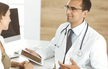 Doktor ve hasta klinikteki masada otururken tıbbi muayene sonuçlarını tartışıyorlar. Genç bir kadının ilaç geçmişini doldurmak için tablet bilgisayar kullanan bir erkek doktor. Tıptaki veriler