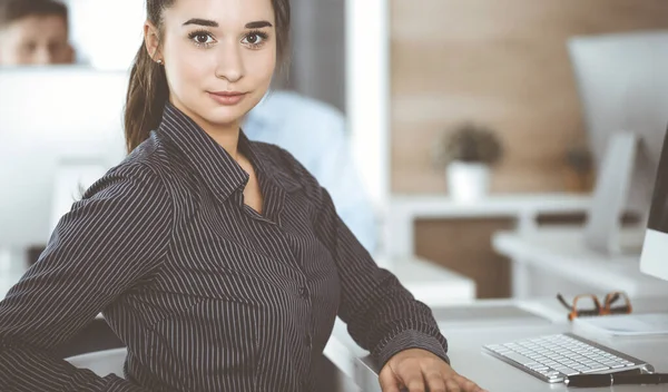 Business woman using computer at workplace in modern office. Brunette secretary or female lawyer looking at the camera and happy smiling. Working for pleasure and success