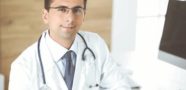 Medico-uomo seduto alla scrivania del suo posto di lavoro e sorridente alla telecamera. Servizio medico perfetto in clinica — Foto Stock