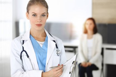 Kadın - doktor, arka plandaki güneşli klinikte tıbbi geçmiş kayıtlarını doldurmak için pano kullanıyor.