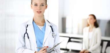 Kadın - doktor, arka plandaki güneşli klinikte tıbbi geçmiş kayıtlarını doldurmak için pano kullanıyor.