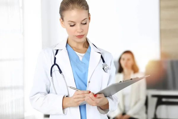 Kvinna - läkare använder Urklipp för att fylla upp medicinering historia rekord form i solig klinik i bakgrunden av patienten — Stockfoto