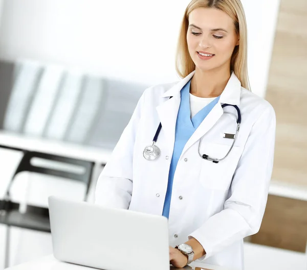 Médica loira está sorrindo ao usar o computador portátil. Mulher-médico no trabalho na clínica animado e feliz de sua profissão. Conceito de medicina — Fotografia de Stock