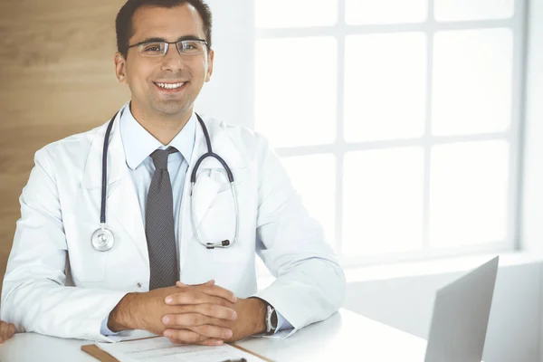 Άνθρωπος-γιατρός κάθεται στο γραφείο στο χώρο εργασίας του και χαμογελά στην κάμερα. Τέλεια ιατρική υπηρεσία στην κλινική — Φωτογραφία Αρχείου
