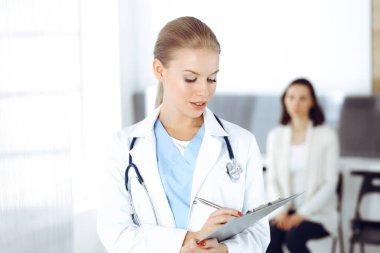 Kadın - doktor hastanın arka planında klinikte tıbbi geçmiş kayıtlarını doldurmak için pano kullanıyor. Tıp konsepti