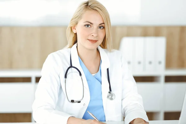 Портрат жінки-лікаря на роботі, сидячи за столом у клініці. Блондинка веселий лікар, що заповнює медичну форму або рецепт — стокове фото