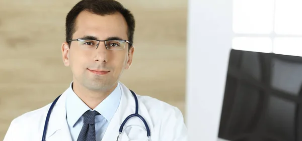 Medico-uomo seduto alla scrivania del suo posto di lavoro e sorridente alla telecamera. Servizio medico perfetto in clinica — Foto Stock