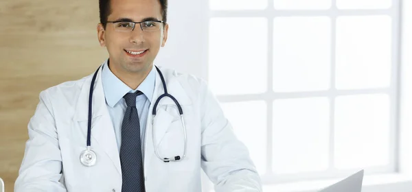 Adam-doktor çalışma yerindeki masada oturuyor ve kameraya gülümsüyor. Klinikte mükemmel bir sağlık hizmeti — Stok fotoğraf