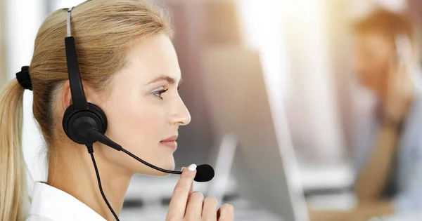Blonďatá obchodnice používající sluchátka pro komunikaci a poradu s lidmi ve slunné kanceláři. Centrum volání — Stock fotografie