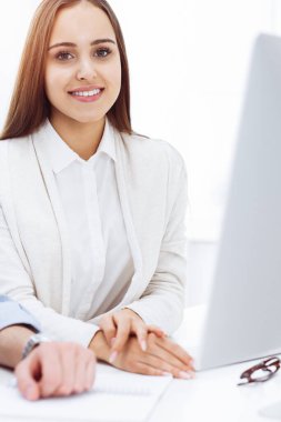 Genç, neşeli bir kadın bilgisayarla masada oturuyor ve güneşli ofisteki kameraya bakıyor. Öğrenci kız ya da iş kadını gibi görünüyor.