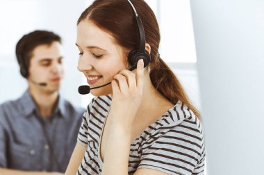 İnternette müşterilerle konuşurken kulaklık ve bilgisayar kullanan sıradan giyinmiş genç bir kadın. Bir grup operatör iş başında. Çağrı merkezi, iş konsepti