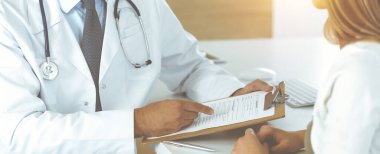 Doktor ve hasta, güneşli klinikteki masada otururken tıbbi muayene sonuçlarını tartışıyorlar. Yakın plan. Erkek doktor pano kullanıyor.