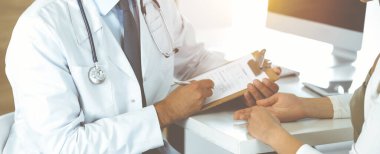 Doktor ve hasta, güneşli klinikteki masada otururken tıbbi muayene sonuçlarını tartışıyorlar. Yakın plan. Erkek doktor pano kullanıyor.