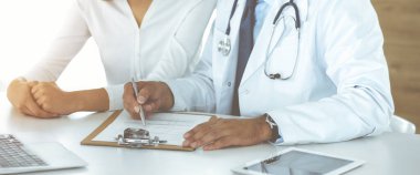 Doktor ve hasta, güneşli klinikteki masada otururken tıbbi muayene sonuçlarını tartışıyorlar. Yakın plan. Erkek doktor pano kullanıyor.