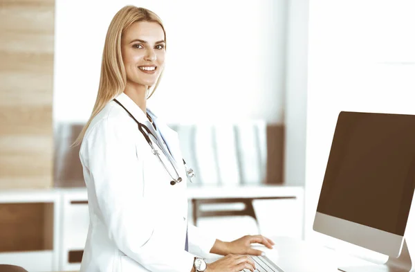 금발의 여성과 의사가 클리닉에서 데스크 탑 컴퓨터로 타이핑을 하고 있습니다. 의사들은 자기 직업에 대해 흥분하고 기뻐하고 있다. 의학적 개념 — 스톡 사진