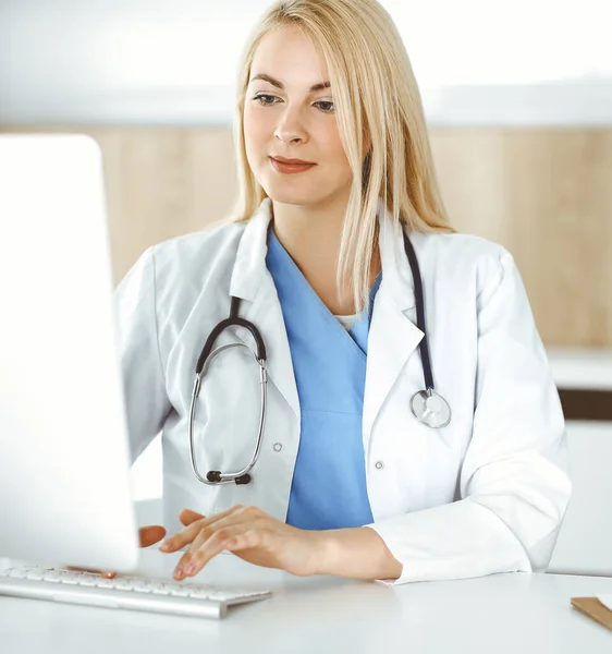 Kobieta-lekarz w pracy podczas siedzenia przy biurku w szpitalu lub klinice. Blondynka wesoły lekarz za pomocą komputera stacjonarnego — Zdjęcie stockowe