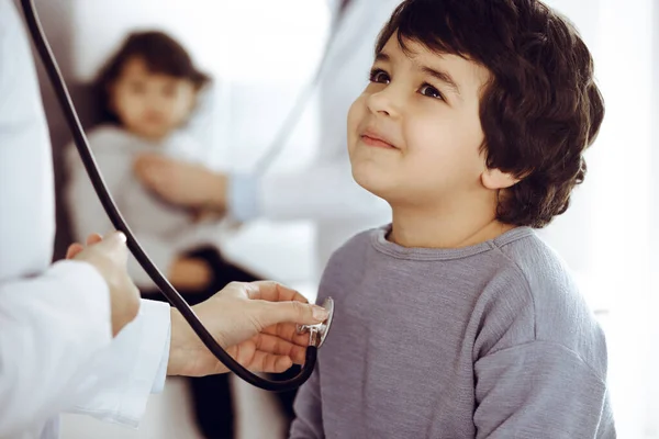 Médico-mujer examinando a un niño paciente por estetoscopio. Lindo chico árabe y su hermano en la cita con el médico. Concepto de medicina —  Fotos de Stock