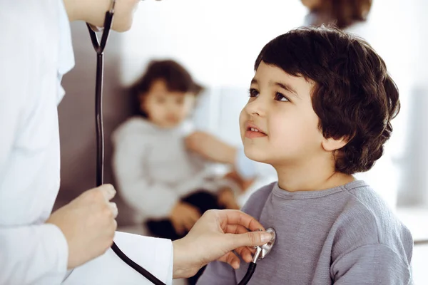 Médico-mulher examinando uma criança paciente por estetoscópio. Rapaz árabe bonito e o irmão na consulta médica. Conceito de medicina — Fotografia de Stock